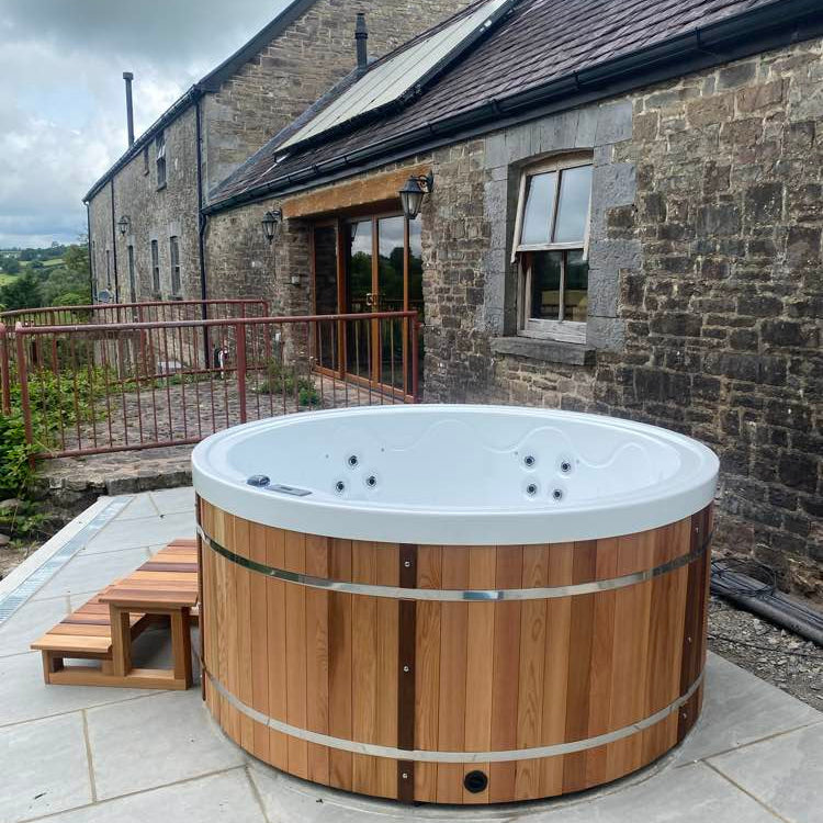 Wooden hot tub on a patio outside a stone building with a cloudy sky.