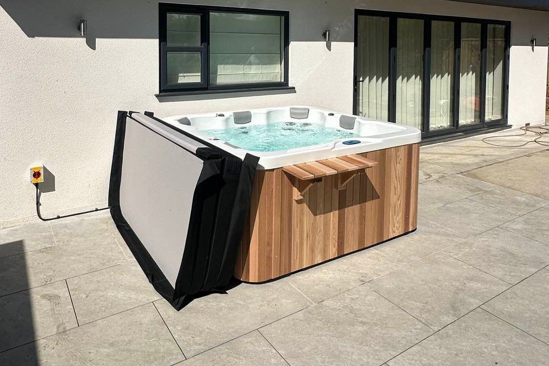 Hot tub with wooden exterior on a patio area in front of a modern house.