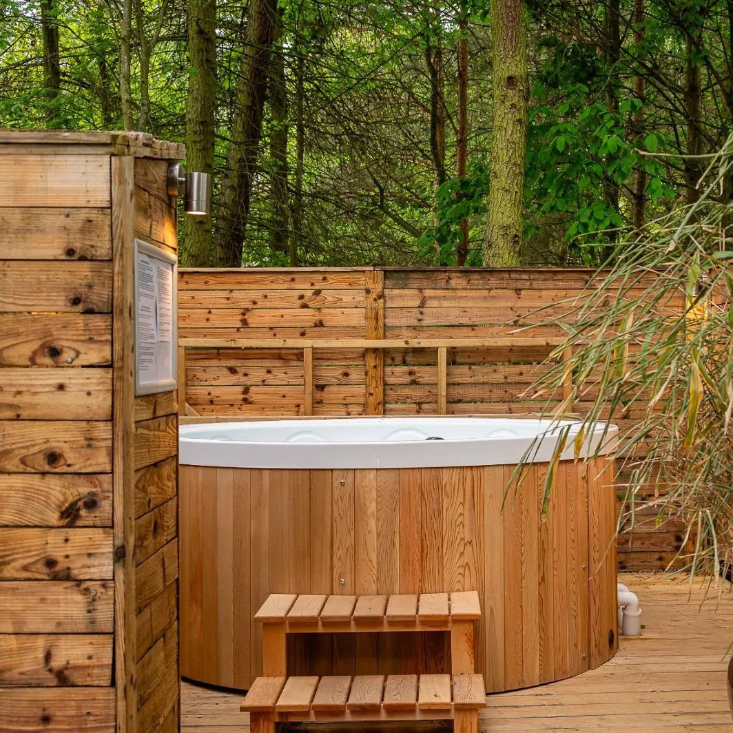 Wooden hot tub in a forest setting with wooden deck and plants.