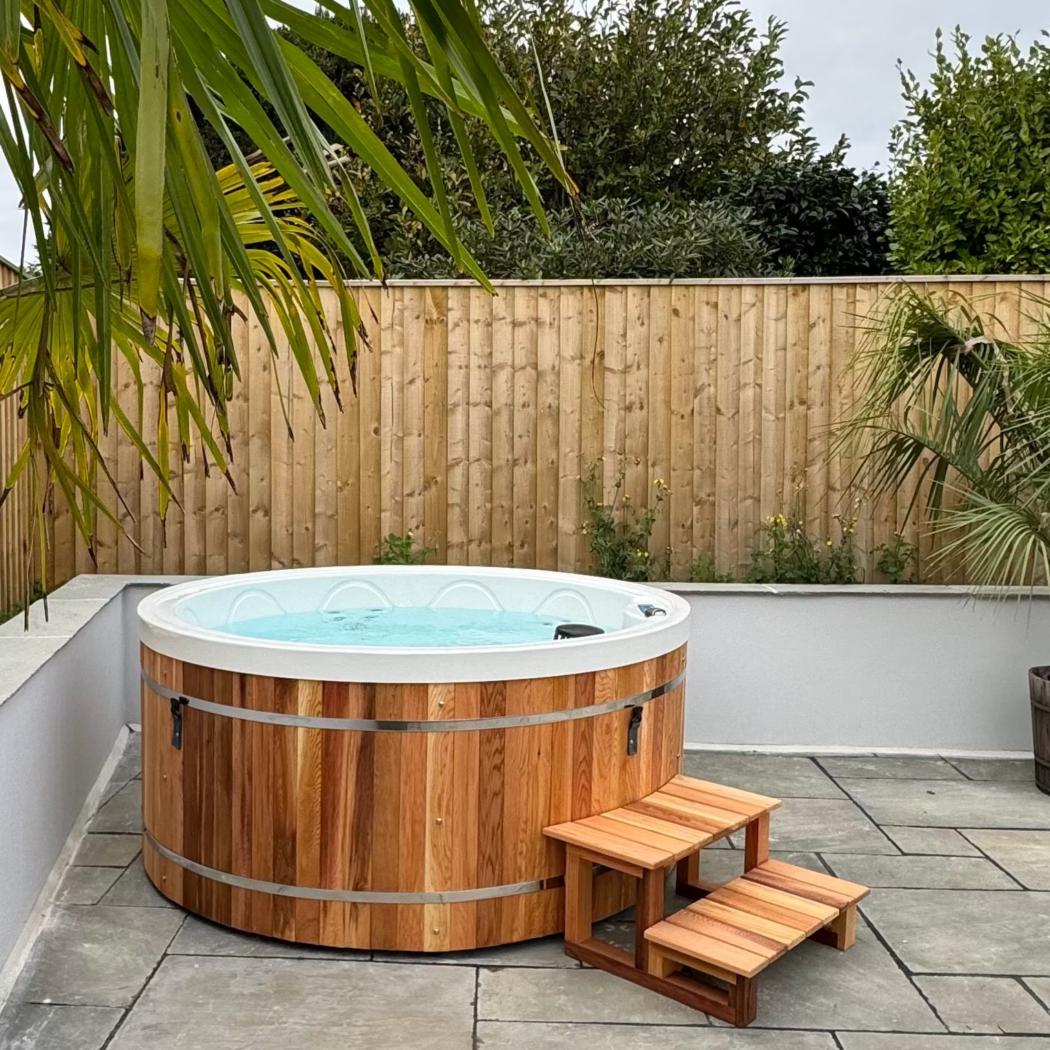 Wooden hot tub with steps on a patio area with plants and wooden fence in the background