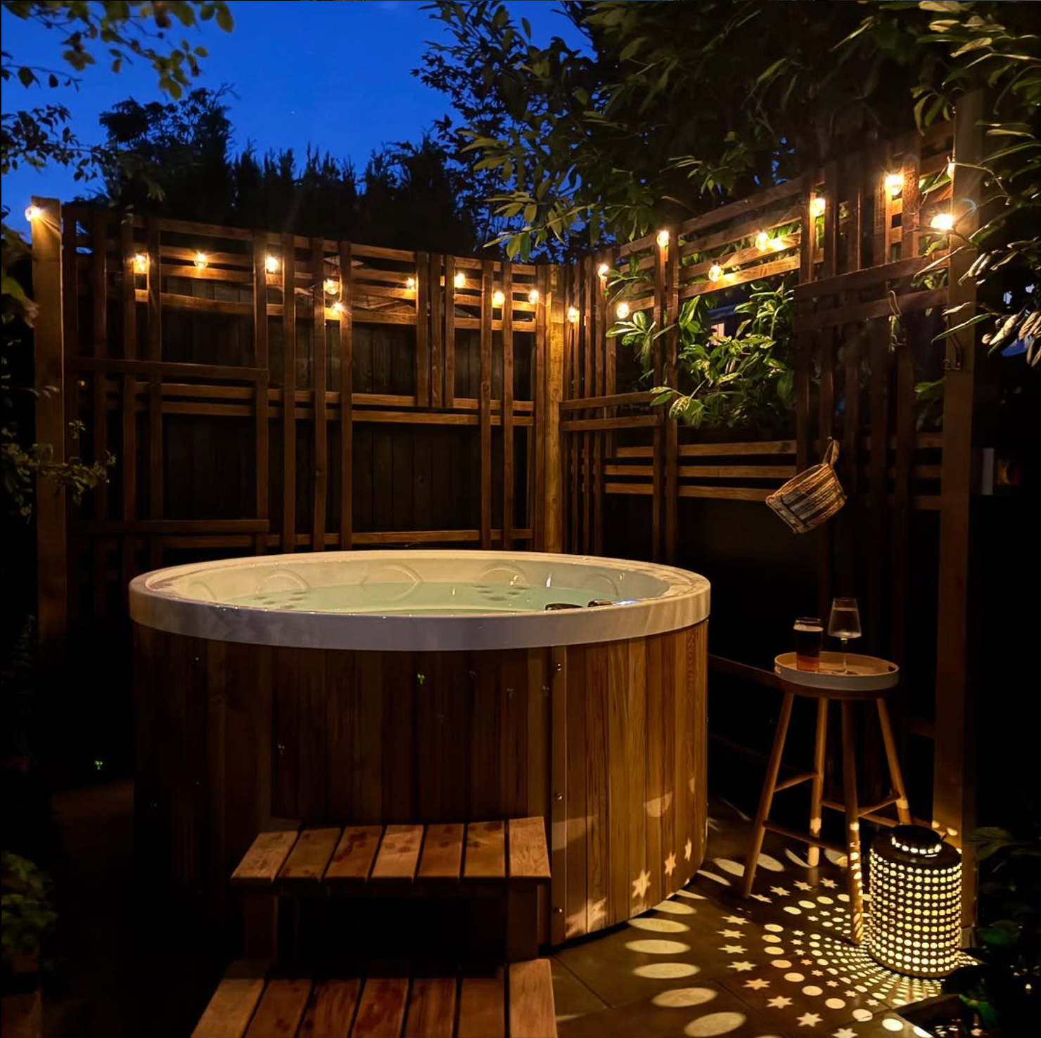 Wooden hot tub in a garden setting with string lights and a stool at night.