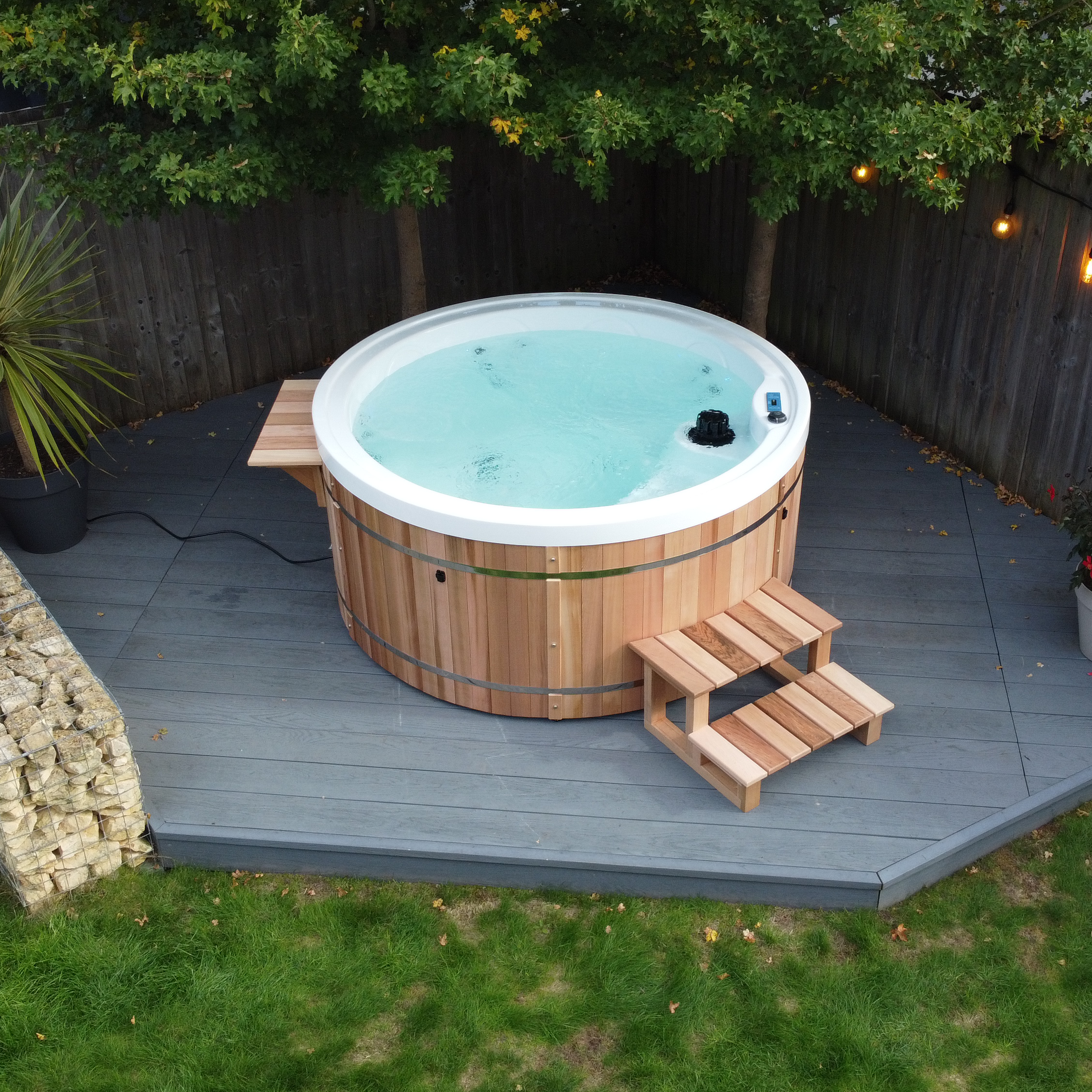 Wooden hot tub on a deck with greenery and lighting in the background