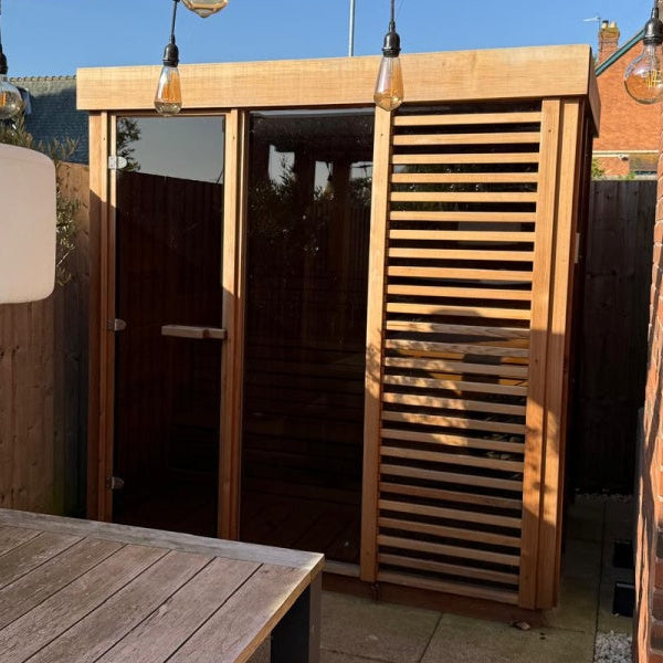 Outdoor sauna with clear glass door and cedar wood walls, featuring a wooden bench and a hanging white light fixture.